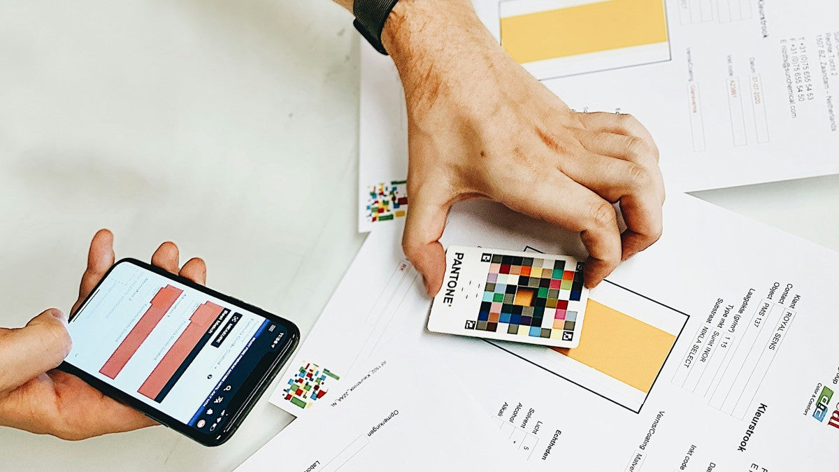 Person holding a smartphone with a design app open, next to color swatches and printed materials on a desk.