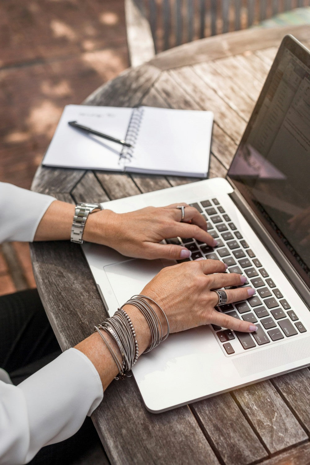 Person typing on a laptop at a wooden table with a notebook and pen.